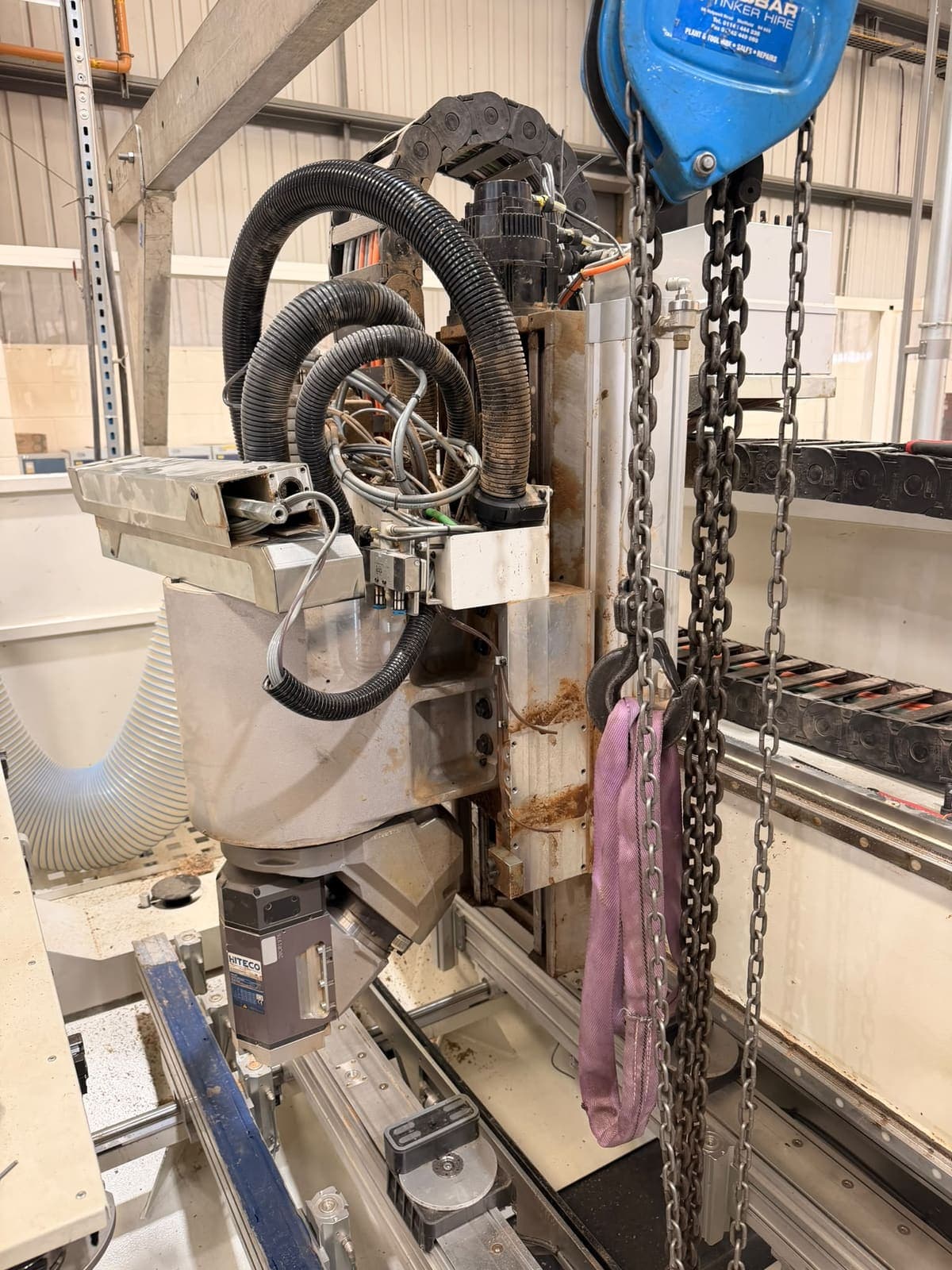 A CNC machining head lifted clear of the machine on a chain hoist, cables and extraction hoses still attached