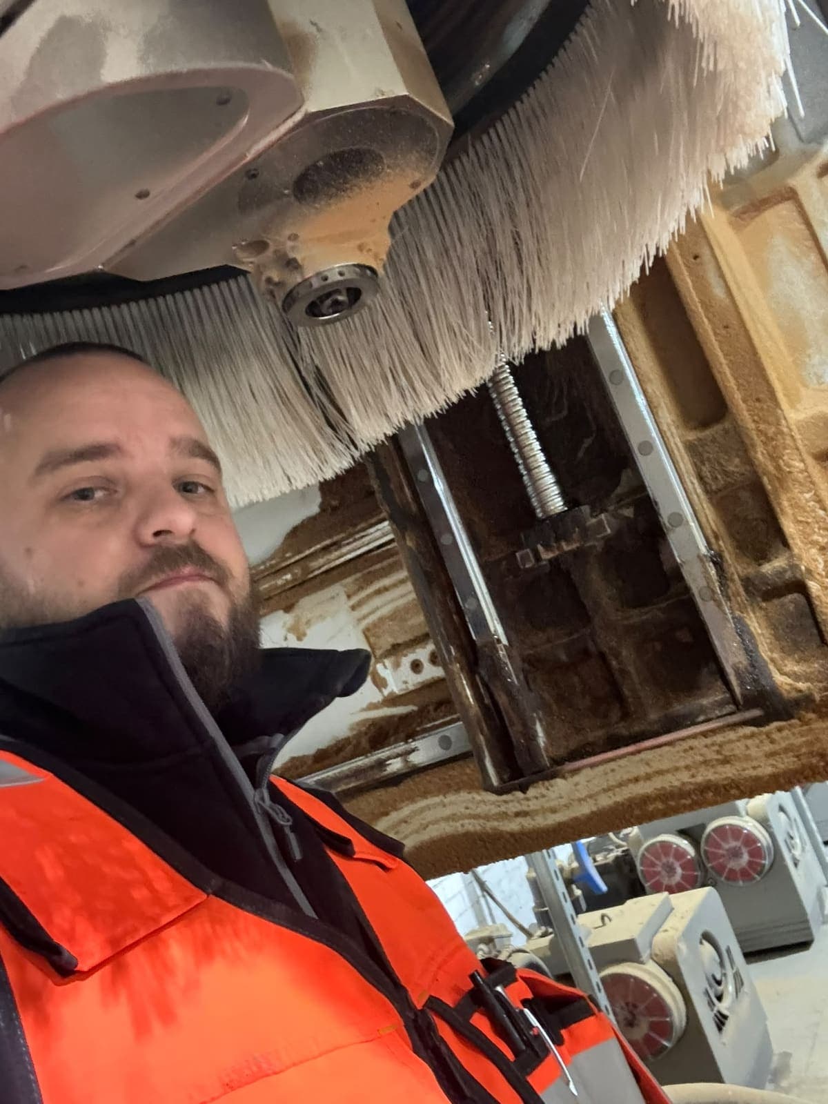 Paul Ashall in an orange hi-vis jacket taking a photo of himself underneath the spindle and dust brushes of a CNC router